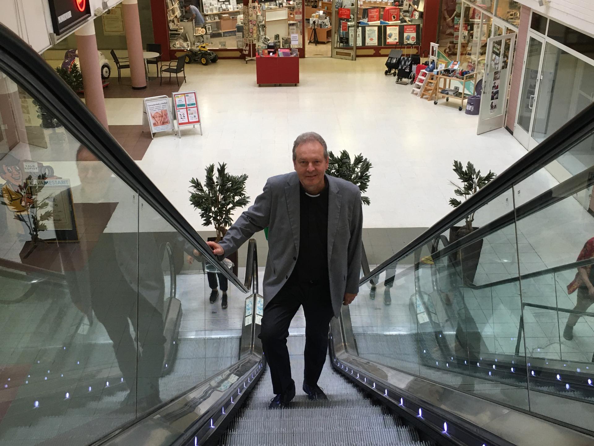 Munkkiniemen seurakunnan seurakuntapastorin sijainen Taneli Hassinen seisoo hymyillen liukuportaissa Munkkivuoren ostarilla